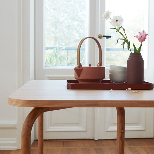 Auf einem hellen Holztisch steht ein rotes terracottafarbenes Tablett mit einer hellroten Teekanne in reduziert geometrischem Stil mit großem Holzgriff. Neben der Kanne stehen weiße Schüsseln und eine dunkelrote Vase mit Blumen.