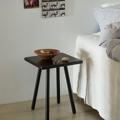Ein schlichter, schwarzer Beistelltisch steht neben einem Bett mit weißer Leinenwäsche.